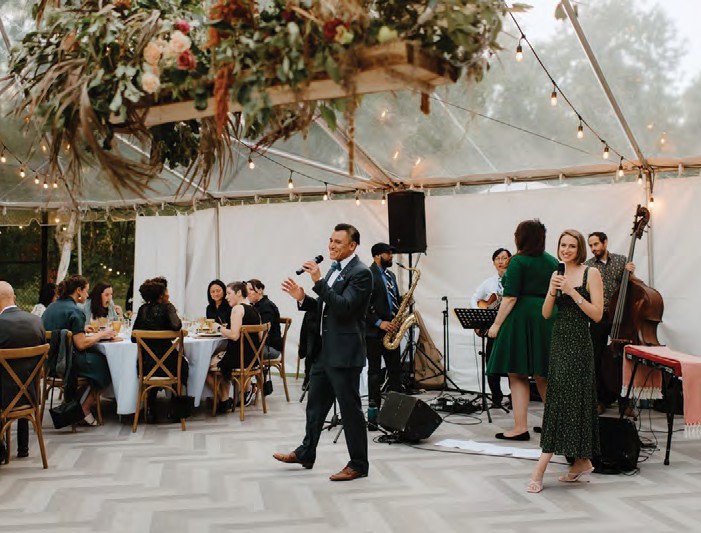Male singer in a suit performs with a live band under string lights at an outdoor reception, while guests sit at tables and enjoy the evening.
