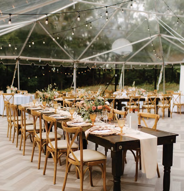 Outdoor clear-tent reception with long tables, wooden chairs, floral centerpieces, and string lights
