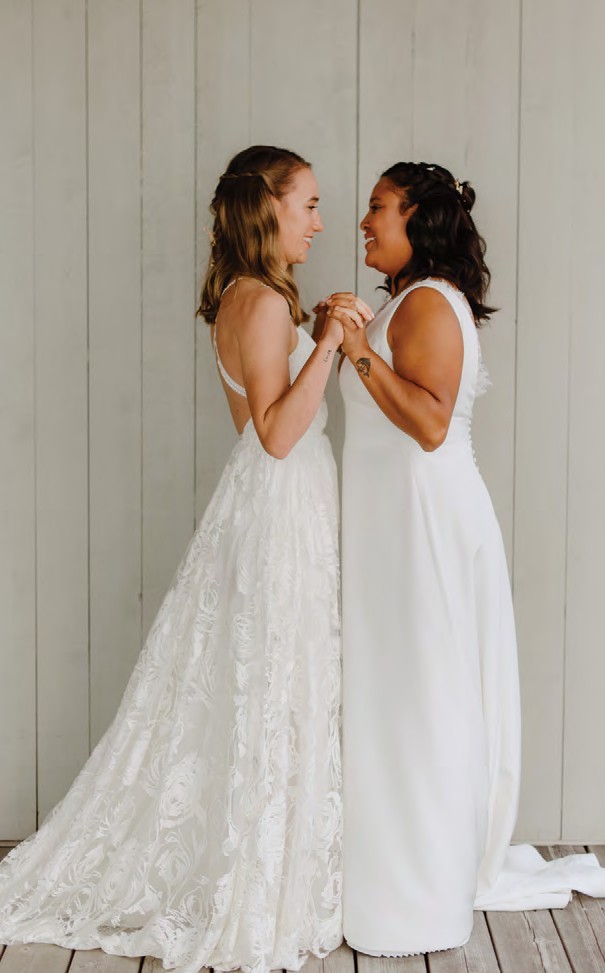 Two brides in wedding dresses face each other, smiling and holding hands.