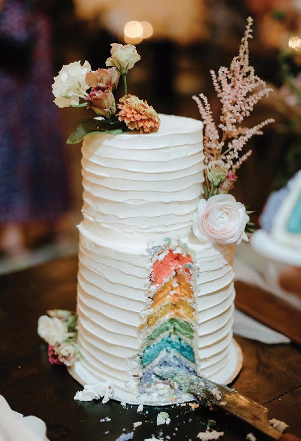 Tall white layered wedding cake with pastel flowers and a rainbow sponge slice exposed on the side in a dramatic reveal.