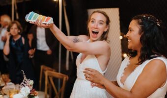 Bride in a white dress holds up a colorful cake slice in celebration, laughing with a guest at a wedding reception.