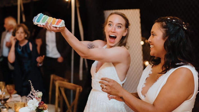 Bride in a white dress holds up a colorful cake slice in celebration, laughing with a guest at a wedding reception.