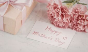 Gift box with pink ribbon, pink carnations, and a 'Happy Mother's Day' note on a marble surface.