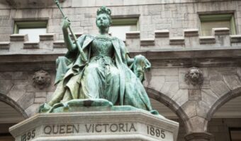 Bronze statue of Queen Victoria seated, holding a scepter, on a pedestal engraved 'QUEEN VICTORIA 1895' with a stone building behind.