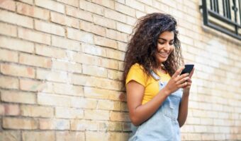 Young woman with curly hair in a yellow shirt and denim overalls checks her smartphone while leaning against a brick wall.