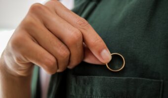 A hand in a dark green shirt holds a gold ring near the shirt pocket, as if placing it inside.