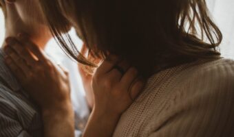 Two people embrace in a close-up, hands resting on each other’s necks and shoulders, warm, intimate lighting.