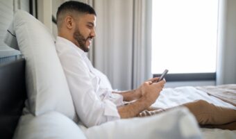 Man relaxing in bed, propped on pillows, looking at a smartphone.