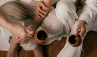 Two people sit on a bed in light-colored clothing, holding brown mugs and brushing hands together in a quiet moment.