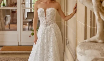 Bride wearing a white lace strapless wedding gown with a fitted bodice and long train, standing by a stone wall indoors.