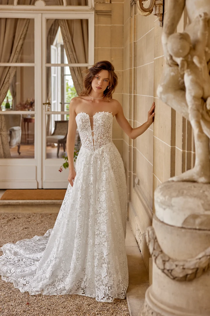 Bride wearing a white lace strapless wedding gown with a fitted bodice and long train, standing by a stone wall indoors.