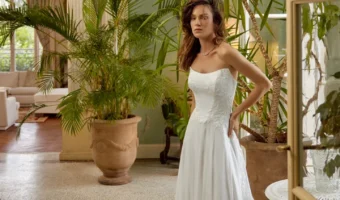 Bride wearing a white strapless wedding gown with lace bodice and long train standing among large potted tropical plants in a sunlit indoor space.