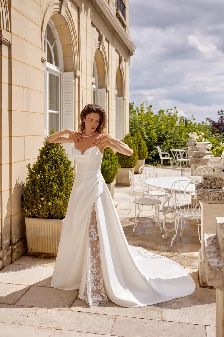 Bride in a strapless white wedding gown adjusting her necklace on a sunlit stone terrace with cream building arches behind her, long train flowing to the side.