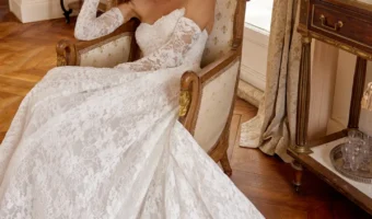Woman wearing an off-shoulder lace wedding gown sits in an antique armchair by a sunlit window, the long lace train spilling onto a wooden floor.