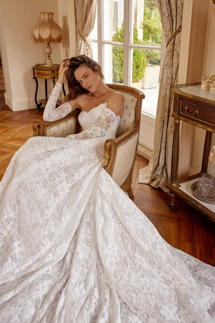 Woman wearing an off-shoulder lace wedding gown sits in an antique armchair by a sunlit window, the long lace train spilling onto a wooden floor.