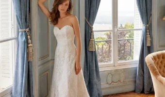 Woman in a strapless lace wedding gown stands by large windows with blue drapes, train flowing on the wooden floor.