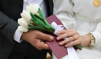 Bride and groom exchanging a small bouquet of white tulips with a burgundy card tied by a white ribbon.