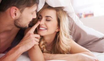 Couple lying in bed under a duvet, the man gently touching the woman's nose as they smile at each other.