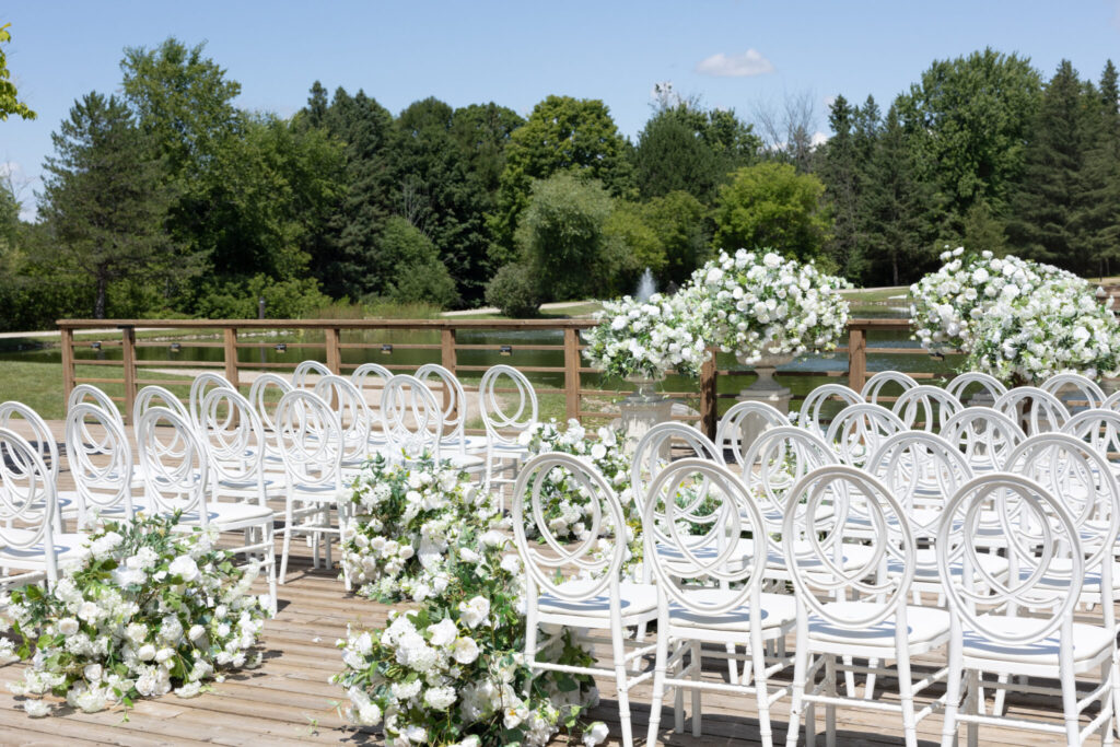 Outdoor wedding ceremony setup on a wooden deck with rows of white decorative chairs and abundant white floral arrangements beside a lake and trees.
