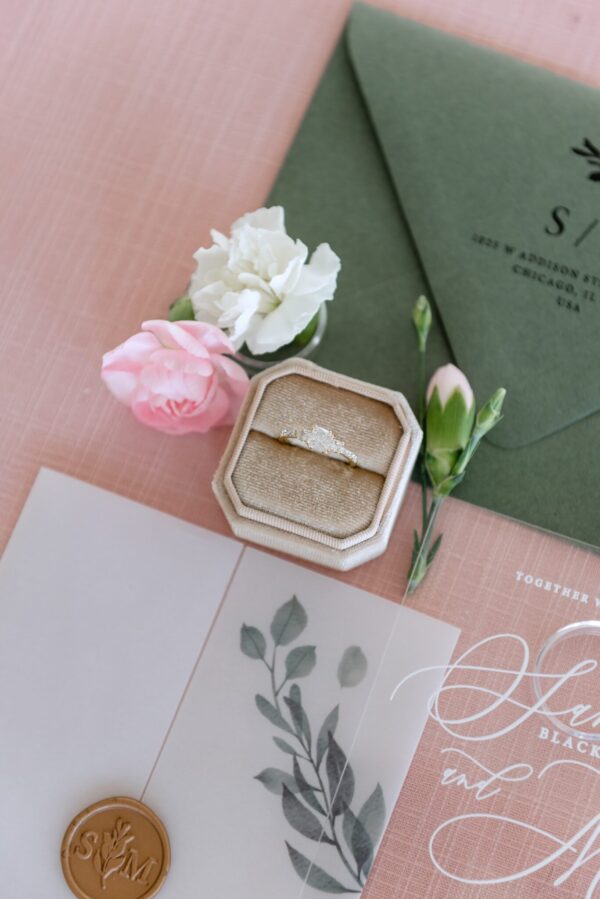 Engagement ring in a beige box with pink and white carnations and a green invitation on a pink backdrop.