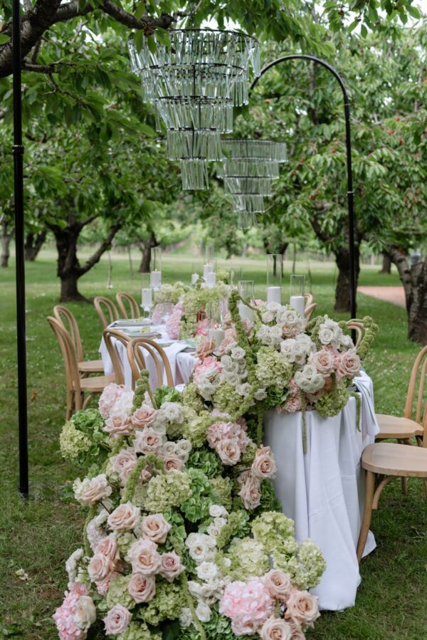 Outdoor garden wedding table set with white linens, pastel pink and white roses and hydrangeas, candle centerpieces, and hanging glass chandeliers above.