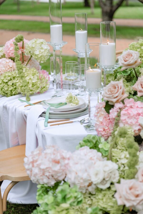 Wedding table centerpiece with white candles in glass cylinders and soft pink/green hydrangea arrangements on a white tablecloth.