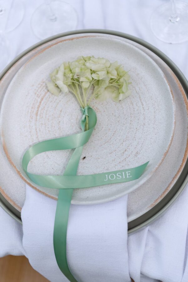 Pale hydrangea boutonniere tied with a mint ribbon placed on stacked ceramic plates in a napkin-wrapped wedding place setting; ribbon reads JOSIE.