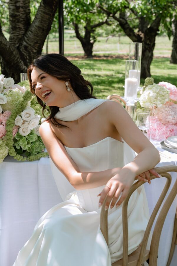 Smiling woman in a white dress sits at a floral outdoor reception table, laughing together with guests.