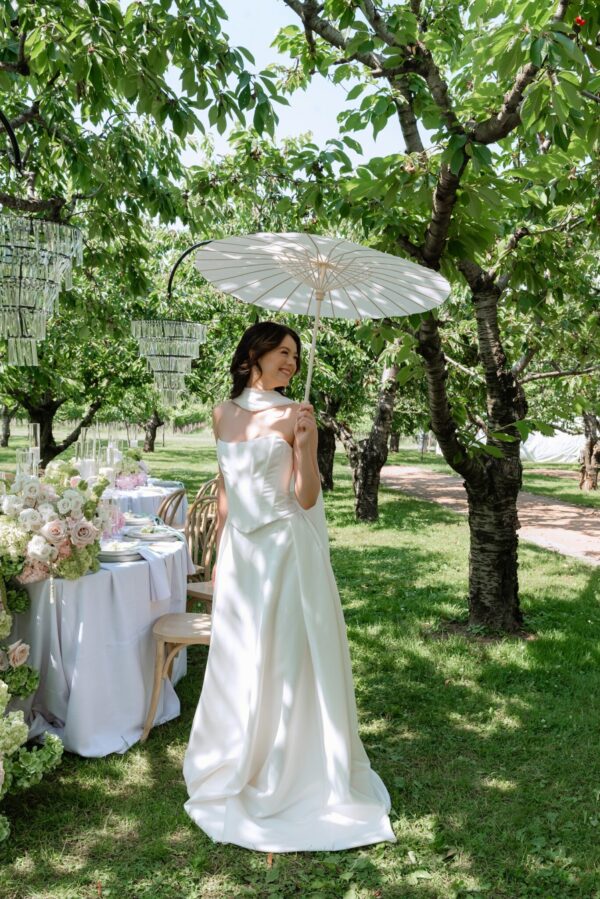 Bride in a white wedding gown holds a parasol under leafy trees at an outdoor reception.