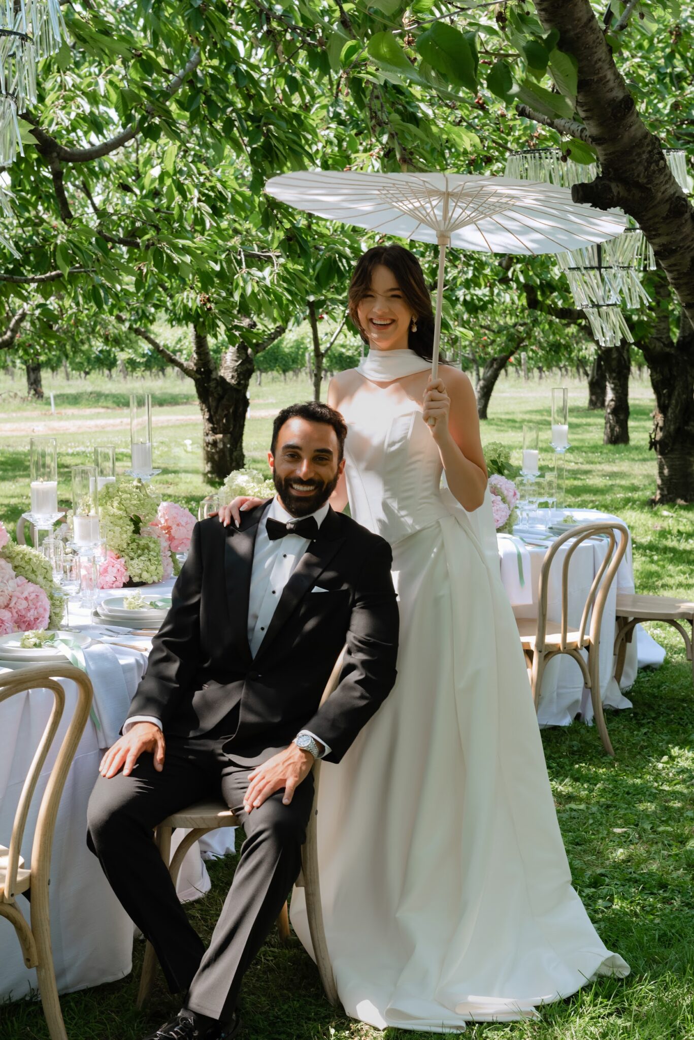 Bride in a white gown stands with a white parasol beside a seated groom in a black tuxedo at an outdoor garden wedding, with a candlelit table and pink flowers nearby.