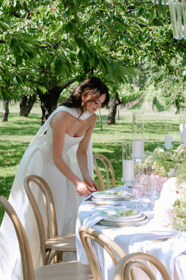 Bride in a white dress arranging an outdoor wedding table beneath green, leafy trees with candles and flowers on the table surface.