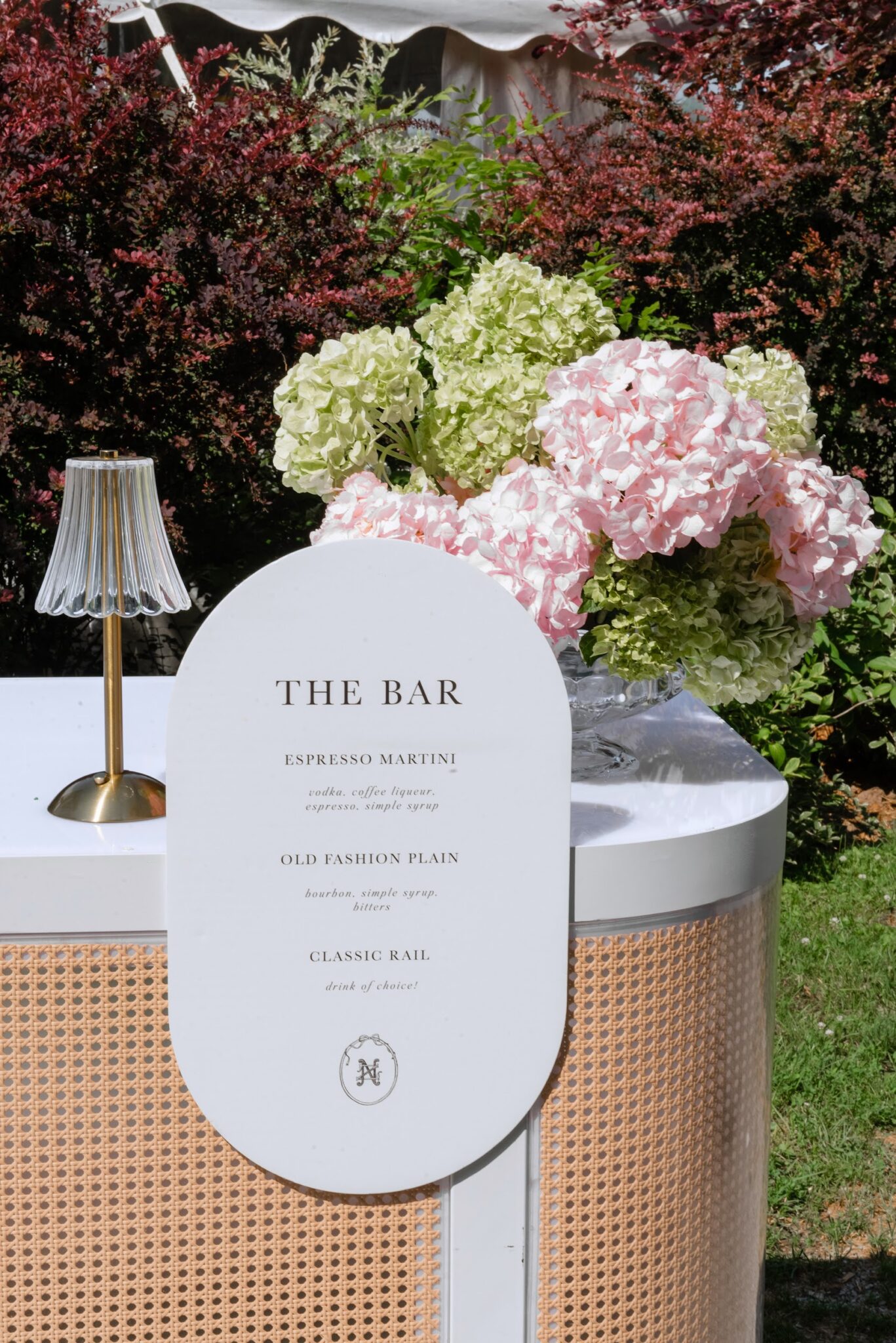 Outdoor event bar setup with a white circular sign reading THE BAR, pale pink and green hydrangea bouquets, and a glass-lamp on a white counter against a shrub backdrop.