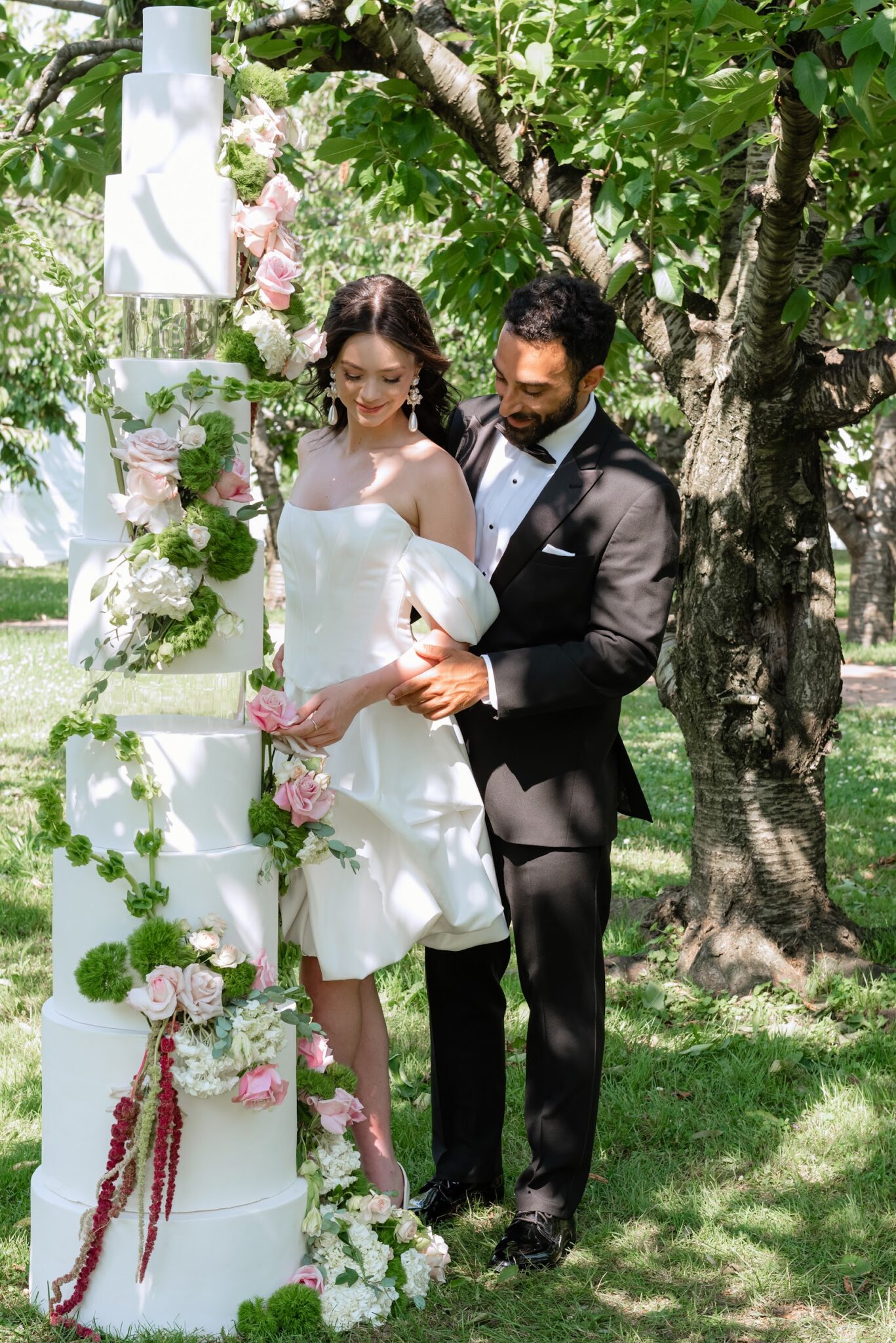 Bride and groom in formal wear posing with a tall white multi-tier wedding cake decorated with pink roses and greenery in a garden.