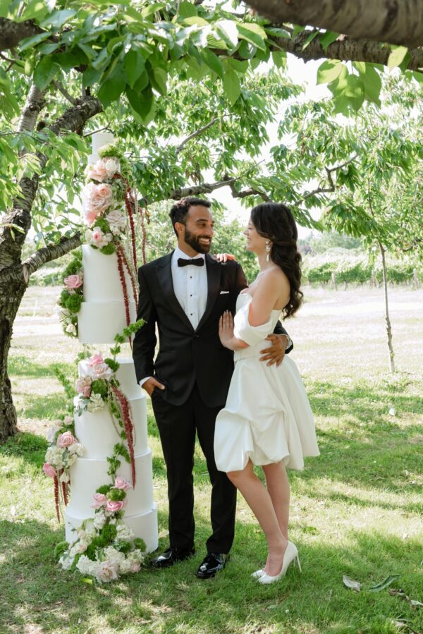 Bride and groom in formal attire pose beside a tall, flower-adorned white wedding cake in an outdoor garden under a leafy tree.