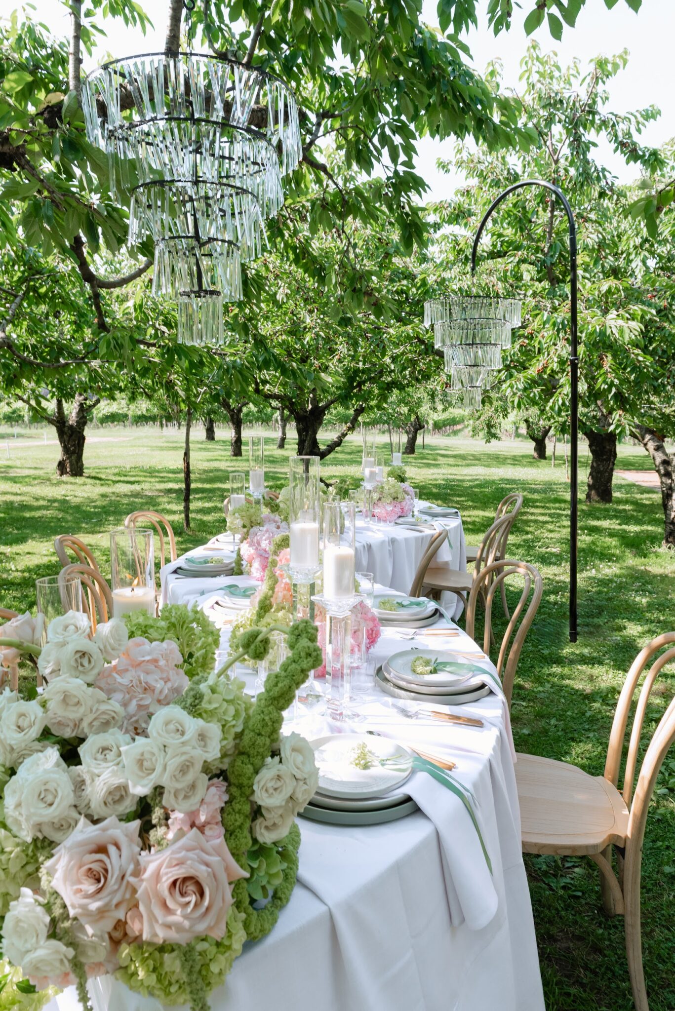 Long white-draped table set for outdoor event with pale pink and white floral centerpieces under hanging crystal chandeliers in a leafy garden.
