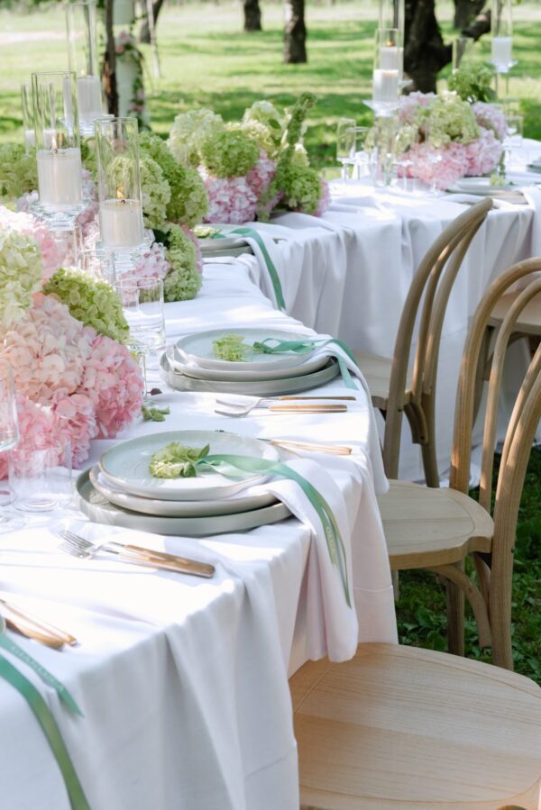 Outdoor wedding table with white linens, pink and green hydrangea centerpieces, glass candleholders, and place settings.