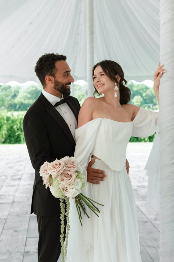 Bride in an off-shoulder white gown holds a blush bouquet while smiling at the groom in a black tuxedo under a white tent.
