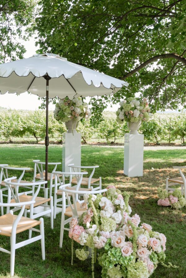 Outdoor wedding ceremony setup with white chairs under a large white parasol and lush greenery and pastel floral arrangements in vases on white pedestals