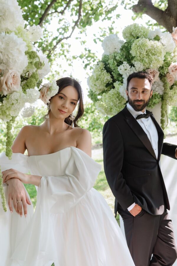 Bride in an off‑the‑shoulder white wedding gown and a groom in a black tuxedo pose under a lush arch of white and pale green flowers.