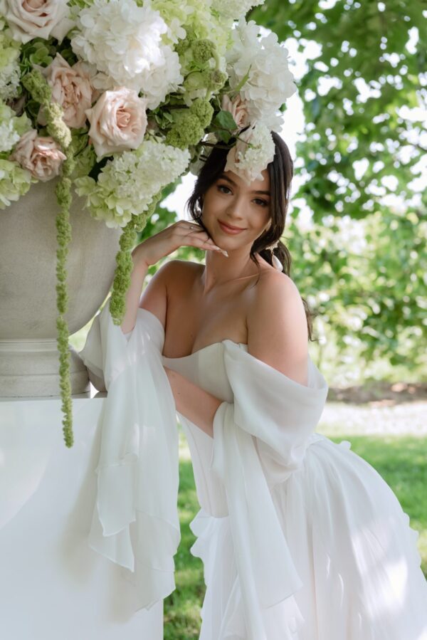 Woman in a white off-shoulder dress posing beside a large floral arrangement of white and pale pink flowers outdoors.