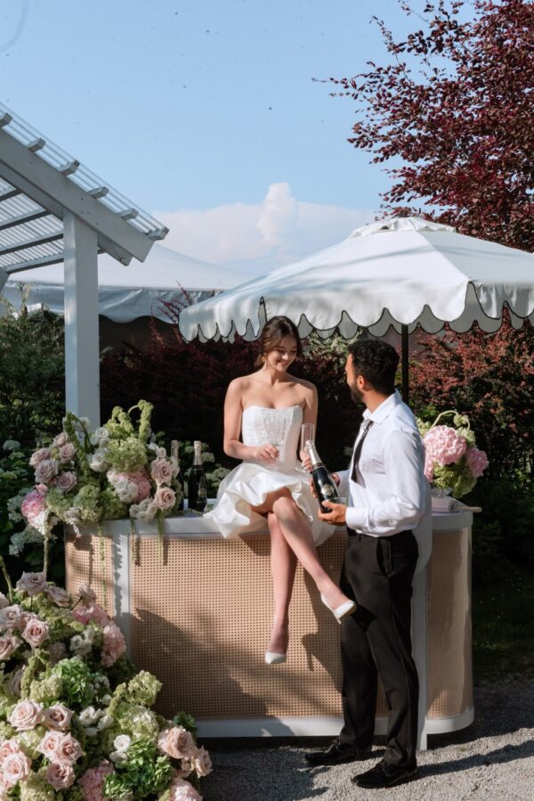Bride in a white dress and groom in a white shirt toast with champagne at a flower-decorated bar under a white canopy garden scene.