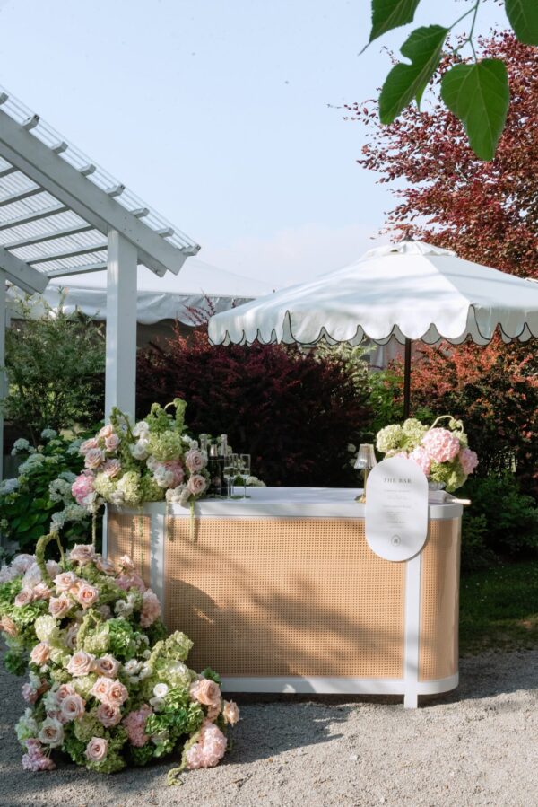 Outdoor bar setup at a garden party, decorated with pale pink and white roses under a white umbrella.
