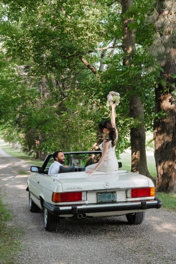 Bride in a white dress stands on a white convertible, bouquet raised high as the groom looks up from the seat, surrounded by trees outdoors.