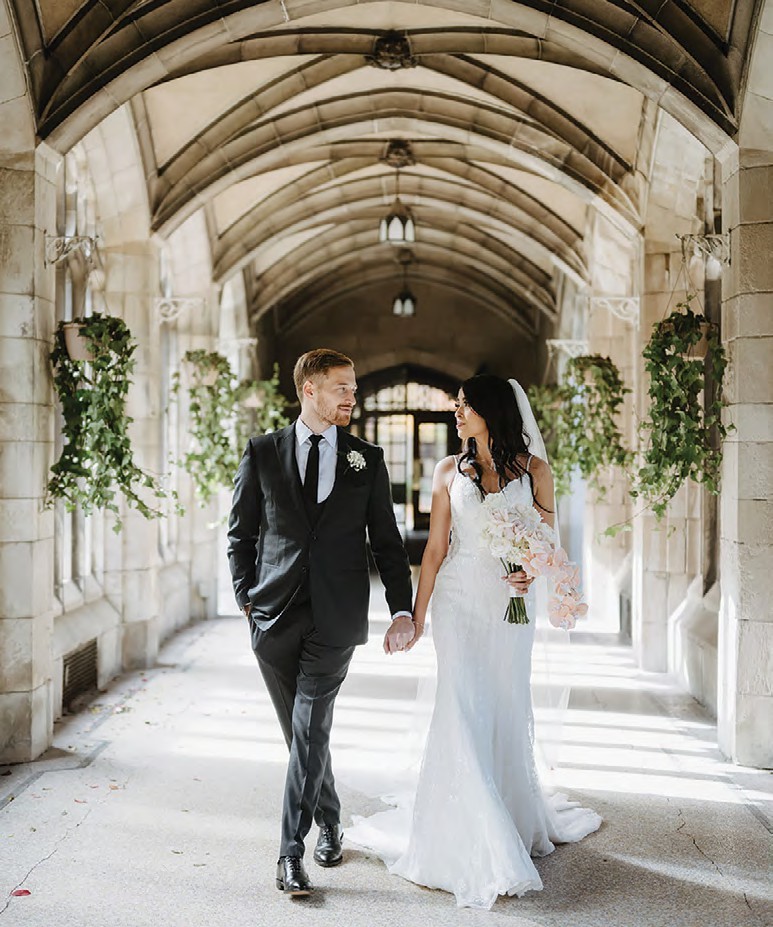 Bride in a white lace wedding gown and veil holding a pale pink bouquet walks hand in hand with a groom in a dark suit under a stone archway lined with greenery.
