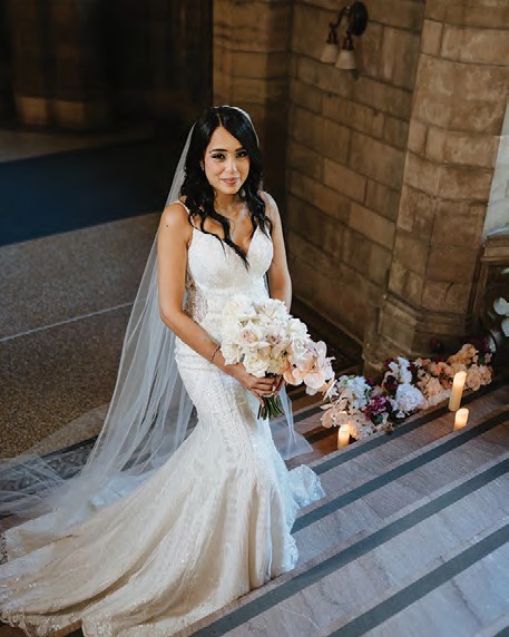 Bride in a white lace wedding gown and veil standing on marble steps holding a pale pink bouquet.