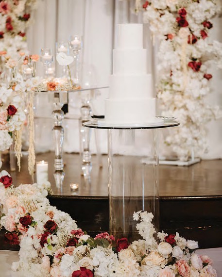 White multi-tier wedding cake on a glass stand, surrounded by ivory and blush floral arrangements.