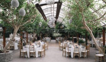Indoor glass conservatory wedding reception with trees, round tables in white linens and gold chairs under a bright skylight.