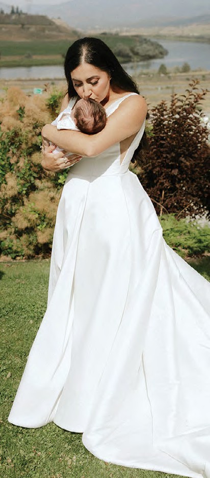 Brunette woman in a white wedding dress cradling a newborn baby outdoors near a garden and lake.
