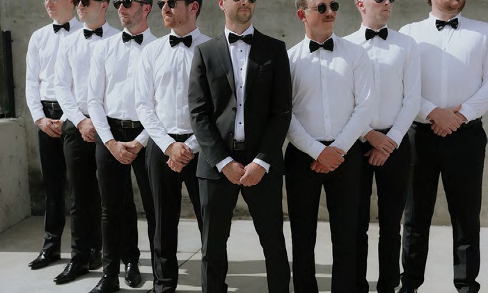 Line of groomsmen in white shirts and black bow ties, centered man in black suit jacket and sunglasses.