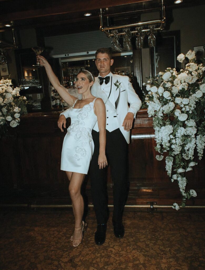 Bride in a white dress raises a glass beside a groom in a white tuxedo at a bar, toasting amid white floral arrangements.